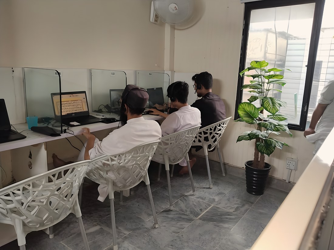 The teaching room in daylight, teachers at their stations with a page of the Quran open on the nearest screen